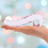 A hand holds the Gläs 6" Lick-It Pink Glass Dildo, a hypoallergenic glass pleasure toy featuring raised knobs, against a softly blurred pastel light background.